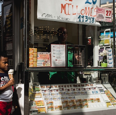 A child holding an ice cream cone stands outside a small kiosk or lottery ticket booth. The booth displays various colorful lottery tickets in the window. There are signs advertising a significant lottery prize amount in Spanish. A person can be seen partially obscured inside the booth, and plants are visible around the area, suggesting an outdoor setting.