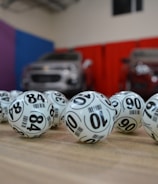 A collection of white lottery balls with black numerals scattered on a wooden surface. Two vehicles are blurred in the background, parked in a room with multicolored walls including teal, purple, blue, and red panels.