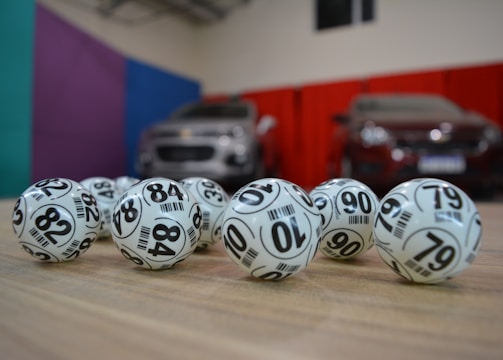 A collection of white lottery balls with black numerals scattered on a wooden surface. Two vehicles are blurred in the background, parked in a room with multicolored walls including teal, purple, blue, and red panels.