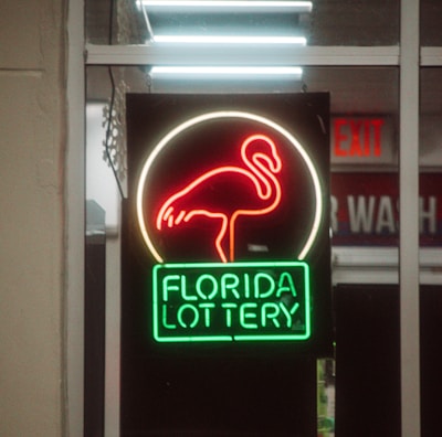 A neon sign depicting a flamingo above the words 'Florida Lottery' is prominently displayed. The sign is illuminated in bright red and green colors, and it is framed by glass panels. An 'EXIT' sign is visible in the background.