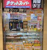 A storefront with various promotional banners and signs in multiple languages advertising ticket exchange services. The counter displays numerous tickets and small items, while the walls are covered with colorful posters and price listings.